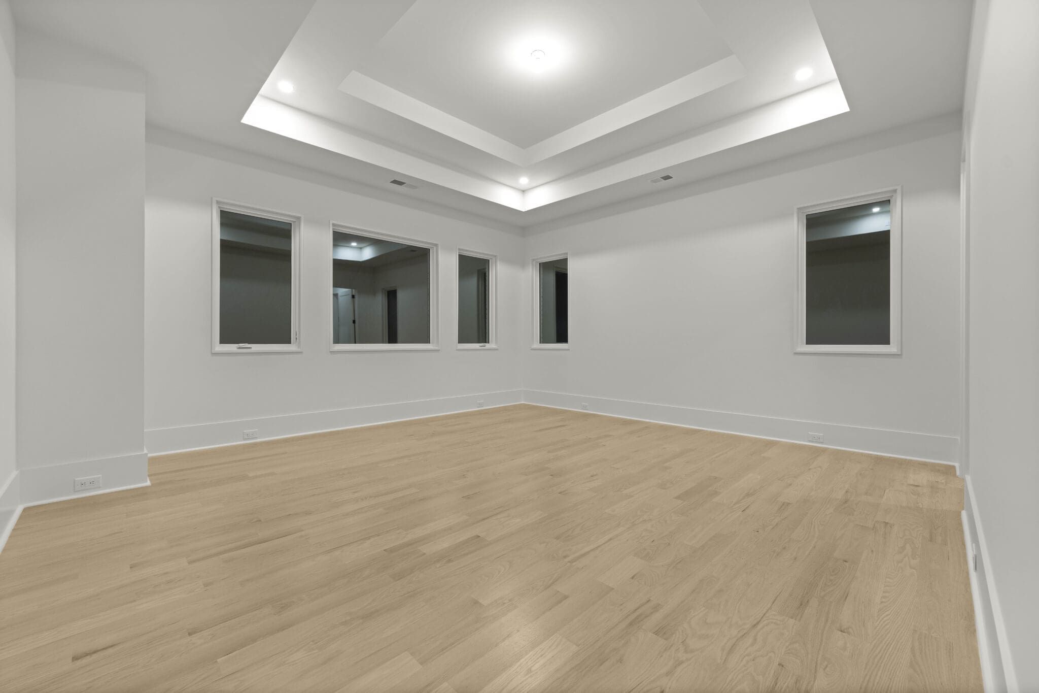 Empty, spacious room with wooden floors, white walls, three windows, and a recessed ceiling with a central light fixture showcases the elegance of luxury home construction.
