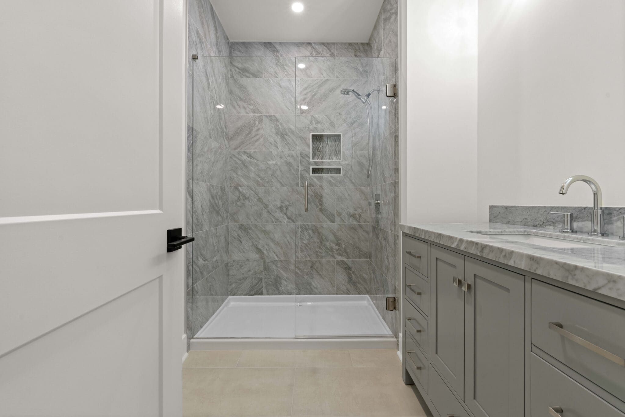A modern bathroom in a luxury home features a glass-door shower with gray tiled walls, a white cabinet with a marble countertop, a silver faucet, beige flooring, and recessed lighting.