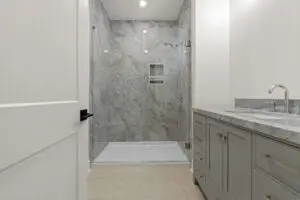 A modern bathroom in a luxury home features a glass-door shower with gray tiled walls, a white cabinet with a marble countertop, a silver faucet, beige flooring, and recessed lighting.