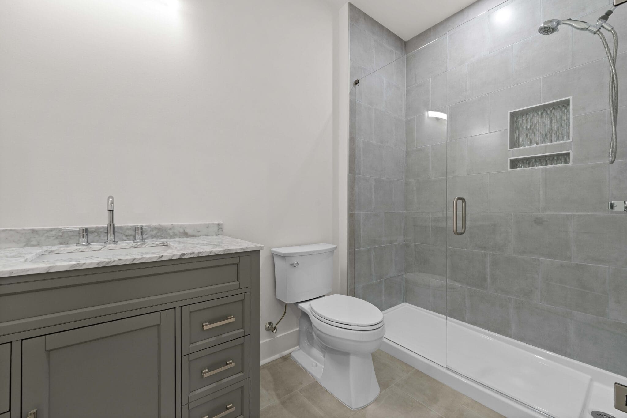 A modern bathroom in a luxury home construction featuring a vanity with a marble countertop, a toilet, and a walk-in shower with grey tiles and a glass door.