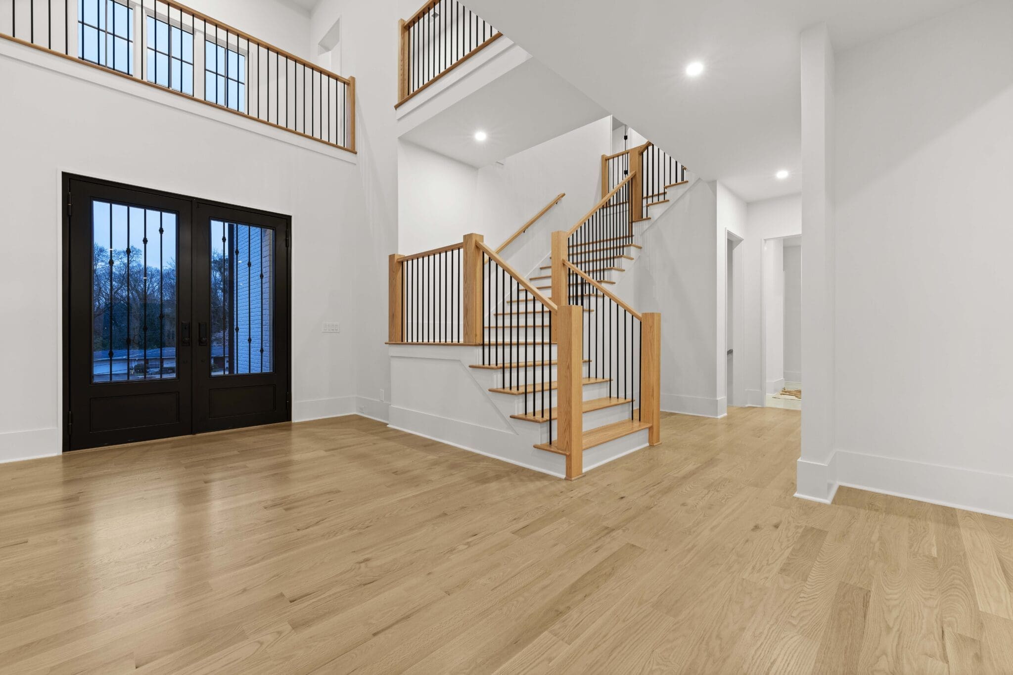 A spacious, modern entryway with light hardwood floors, white walls, and a wooden staircase leading to an upper level. Black double doors with glass panels are on the left, and recessed lights are on the ceiling, showcasing luxury home construction at its finest.