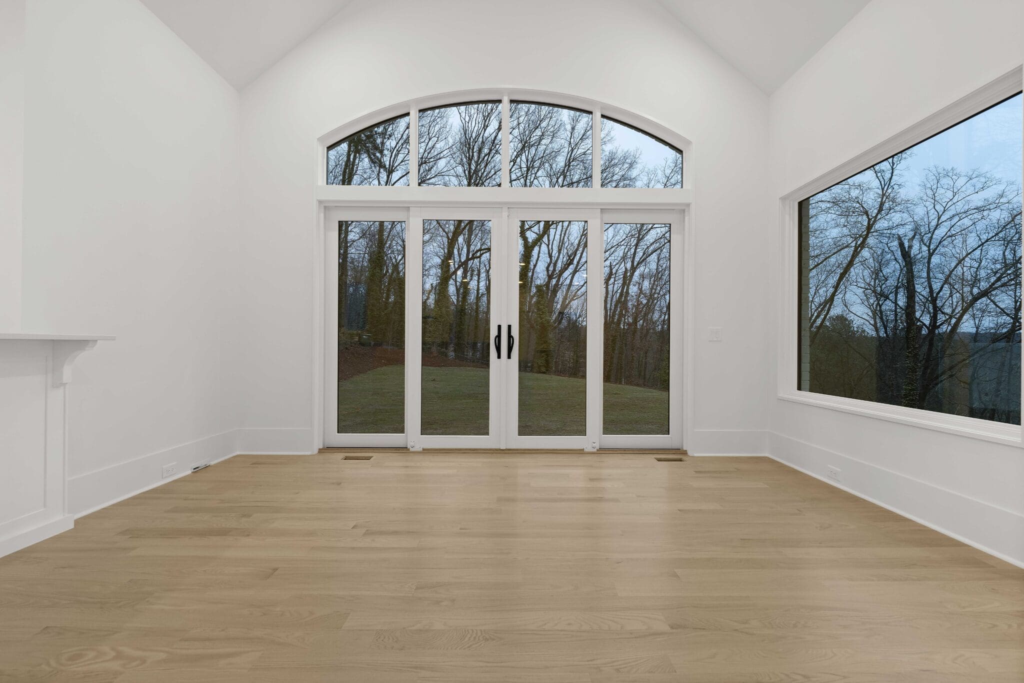     An empty room with light wooden floors, white walls, and a high ceiling exudes the essence of luxury home construction. It features large windows and glass doors overlooking trees and an inviting outdoor area.