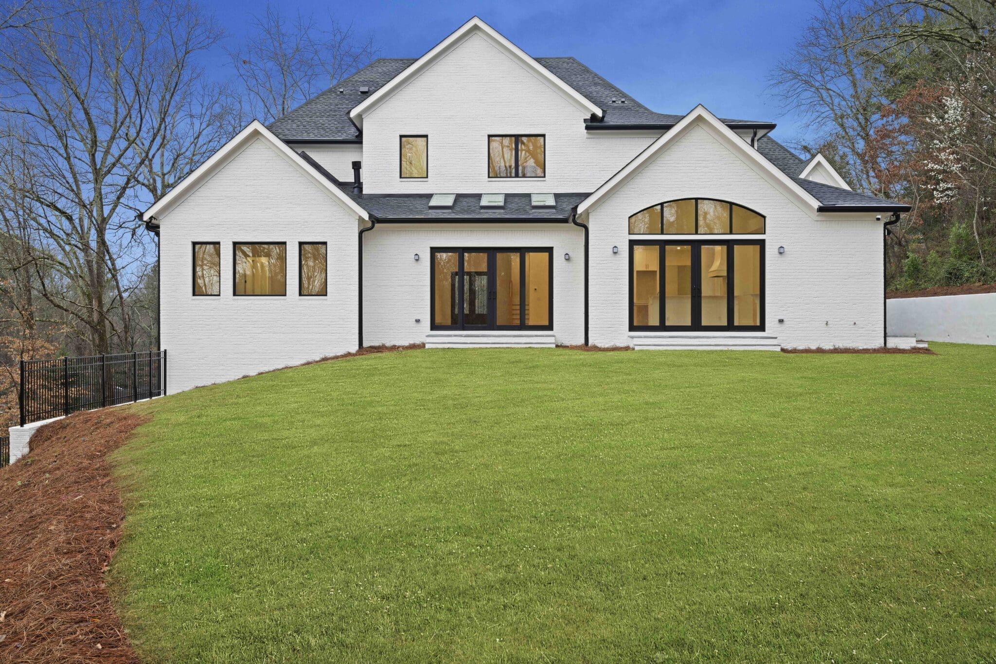 A modern white two-story house with large windows, sloped dark roof, and a spacious green lawn in the foreground showcases luxury home construction. Trees and a cement wall can be seen in the background.
