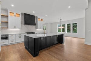 A modern kitchen with a large black island, white cabinets, stainless steel appliances, a black range hood, a tile backsplash, and wooden flooring. Large windows and a dining area are in the background.