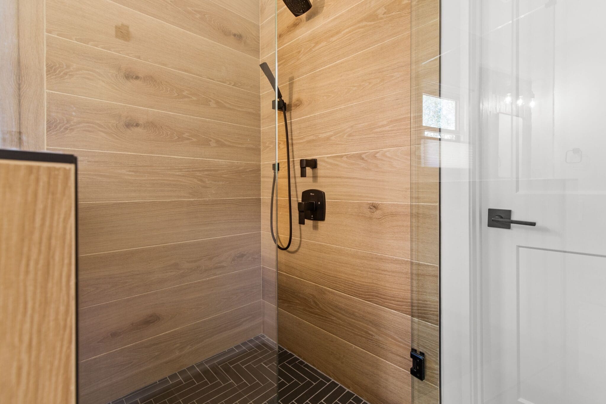 Modern shower with wooden wall panels, a glass door, and a black showerhead and control system. The floor features dark tiles arranged in a herringbone pattern, epitomizing luxury home construction.