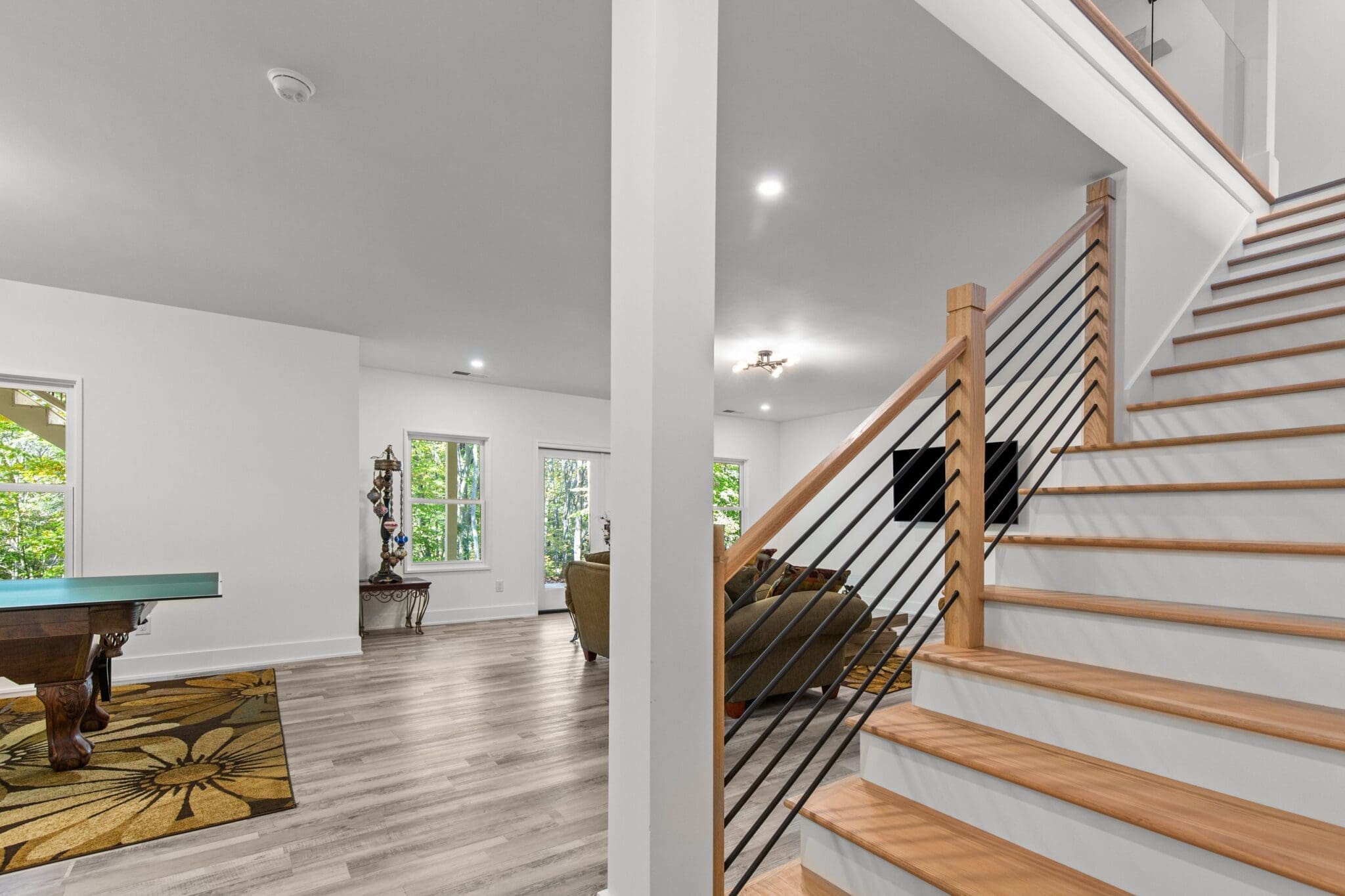 A modern interior, courtesy of luxury home construction, features light wood floors, a staircase with black railings, a living area with a sofa, a wall-mounted TV, and large windows showcasing green foliage outside.