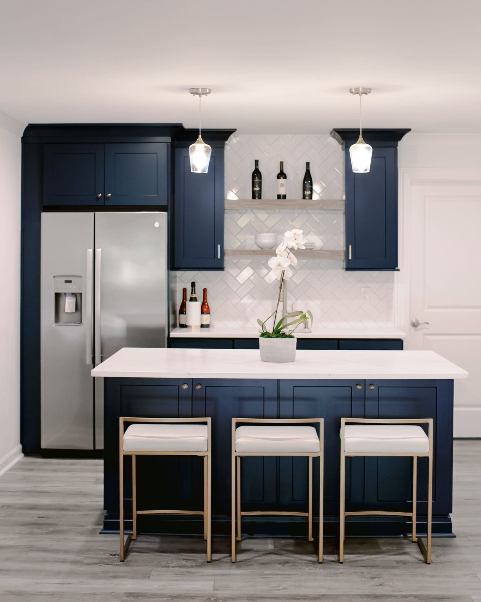 Modern kitchen with navy blue cabinets, a stainless steel refrigerator, white countertop island, and three white bar stools. Two pendant lights hang above the island, and a potted orchid sits on top—a true testament to luxury home construction.