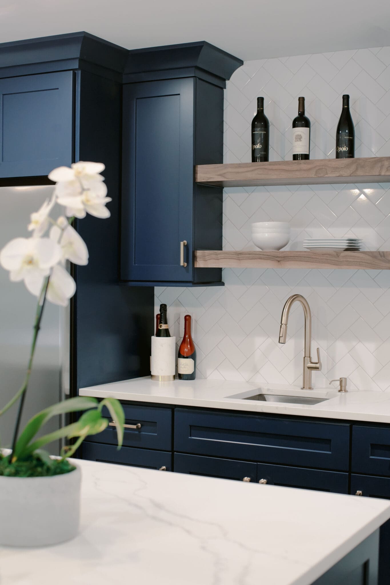 A modern kitchen in a luxury home construction showcases navy blue cabinets, wooden shelves adorned with bottles and bowls, a pristine white countertop, and an elegant orchid in the foreground.
