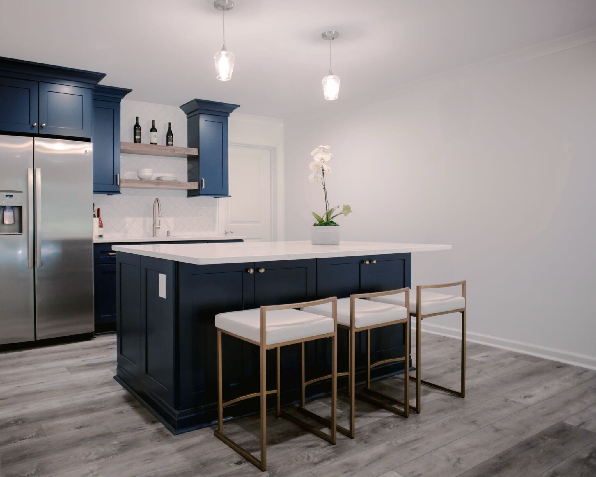 Modern kitchen with blue cabinets, stainless steel appliances, and a white island countertop. Three barstools are positioned at the island, adding to the charm of this luxury home construction. A potted plant decorates the surface, bringing a touch of nature indoors.