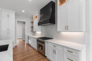 A spacious modern kitchen with white cabinets, stainless steel gas range and hood, hexagonal tile backsplash, wooden floor, and under-cabinet lighting. Open shelves and marble countertops are present.