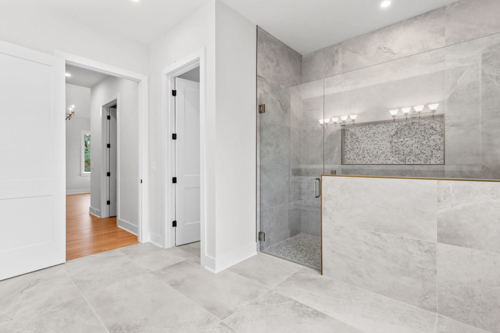 A modern bathroom with a large tiled walk-in shower, glass door, and a doorway leading to a hall with wooden floors.