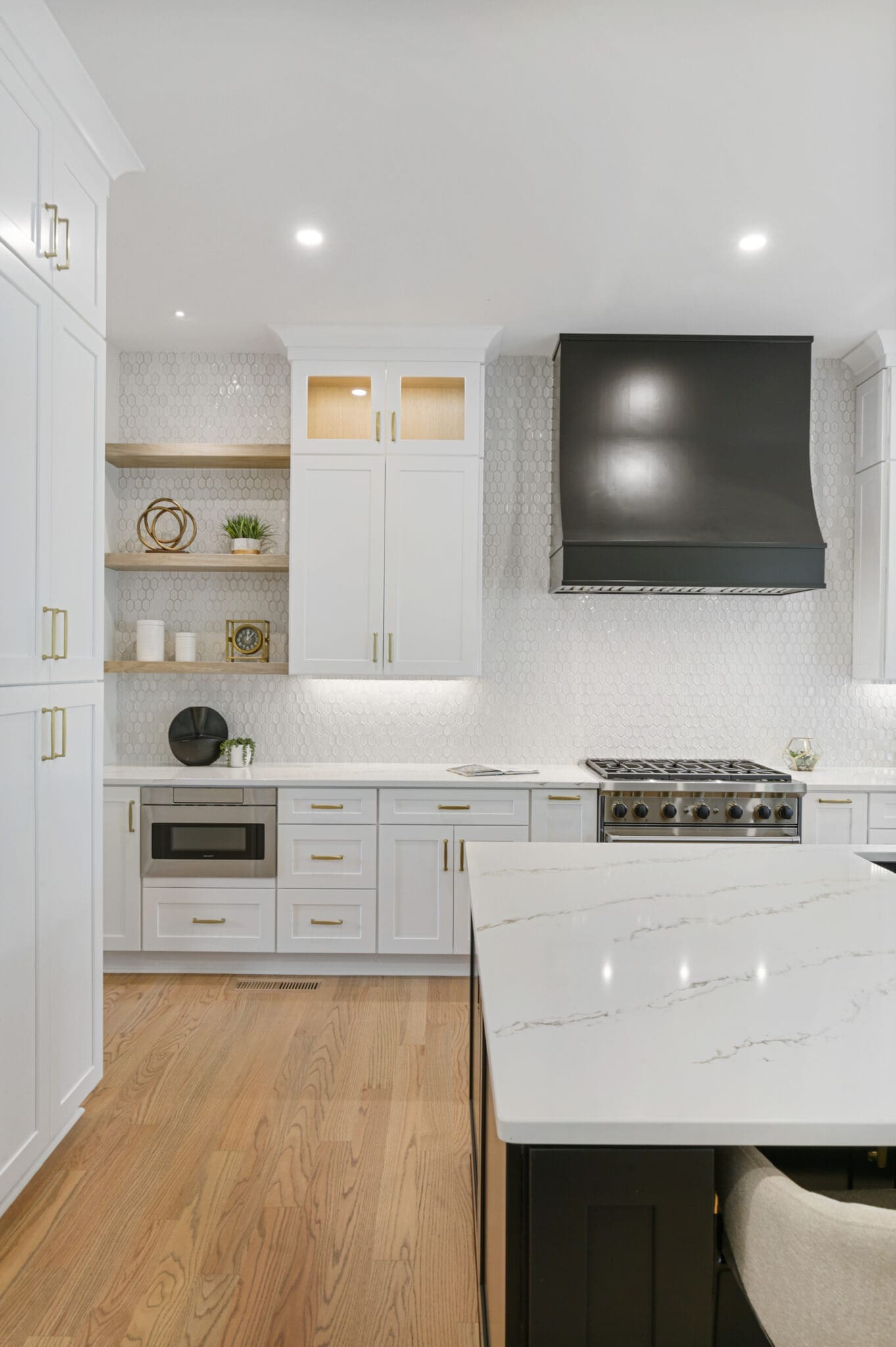 Modern kitchen with white cabinetry, black range hood, stainless steel appliances, and a marble island countertop. Wooden shelves feature minimalistic decor. Light wood flooring completes the space.