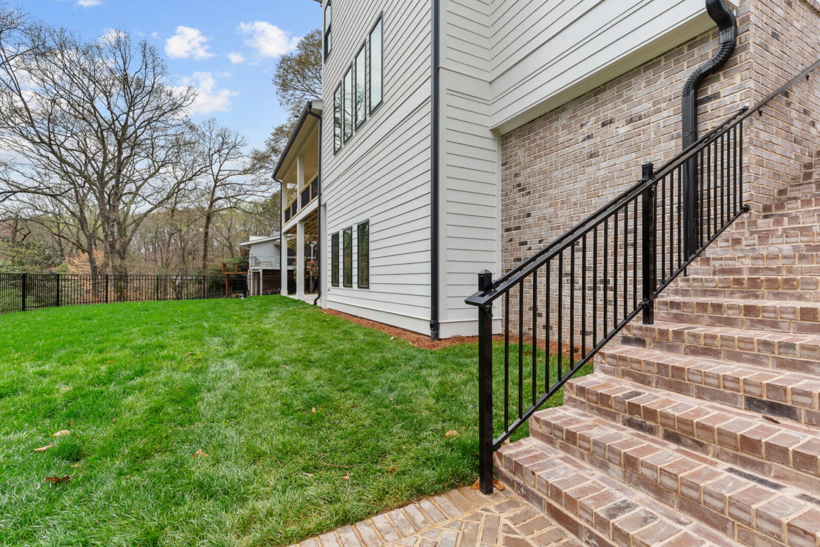 A side view of a modern house with light-colored horizontal siding and brick accents, featuring a brick staircase with a black metal railing, adjacent to a freshly mowed green lawn.