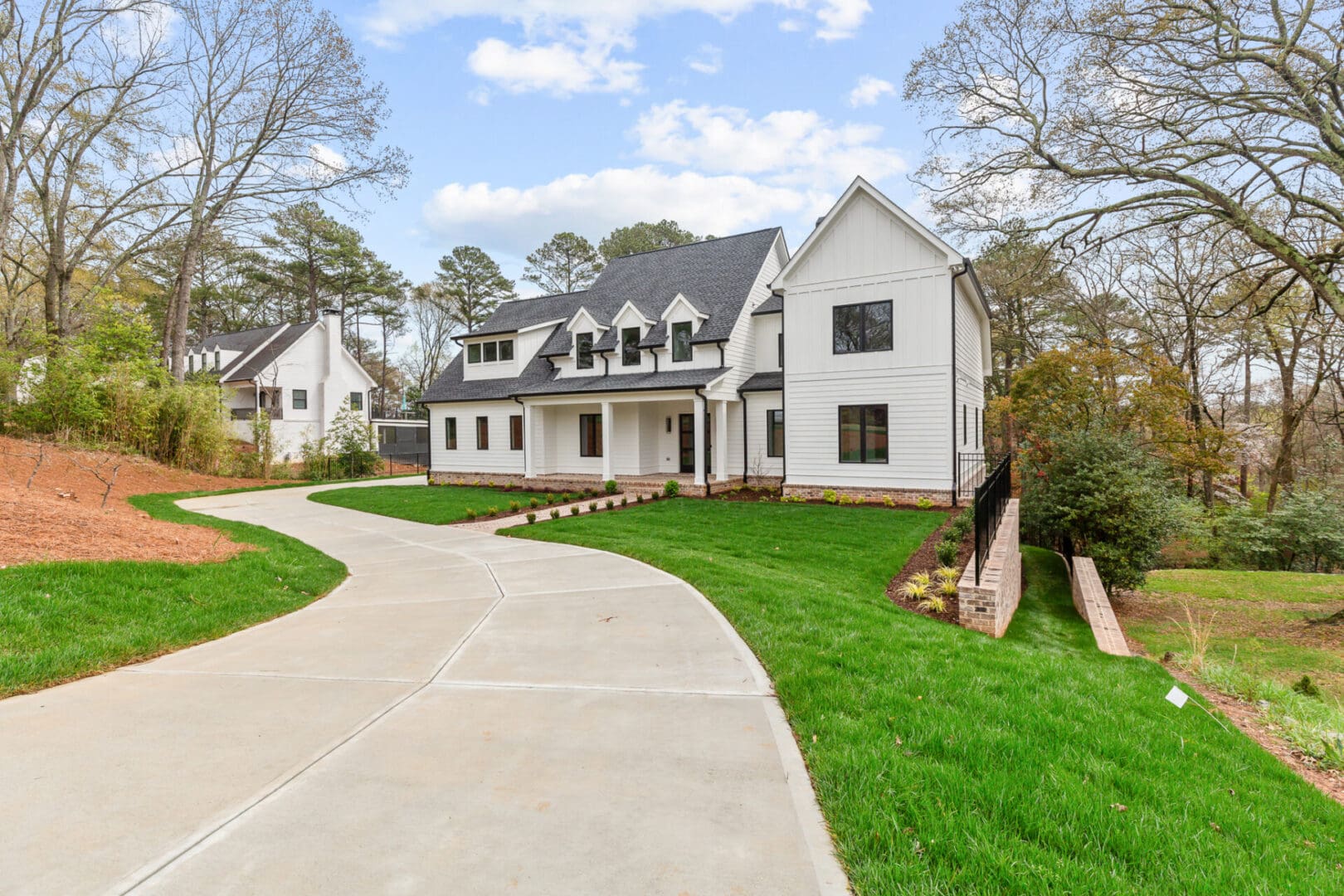 A white, two-story house with dark roof shingles, expansive lawn, and a curved driveway, set amidst trees under a partly cloudy sky.