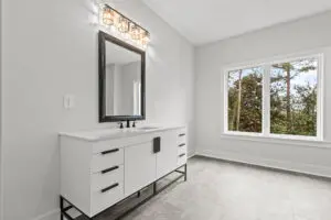 A modern bathroom featuring a white vanity with a black-framed mirror, black fixtures, and a three-light sconce above. A large window offers a view of trees and natural light. Gray tiled floor.