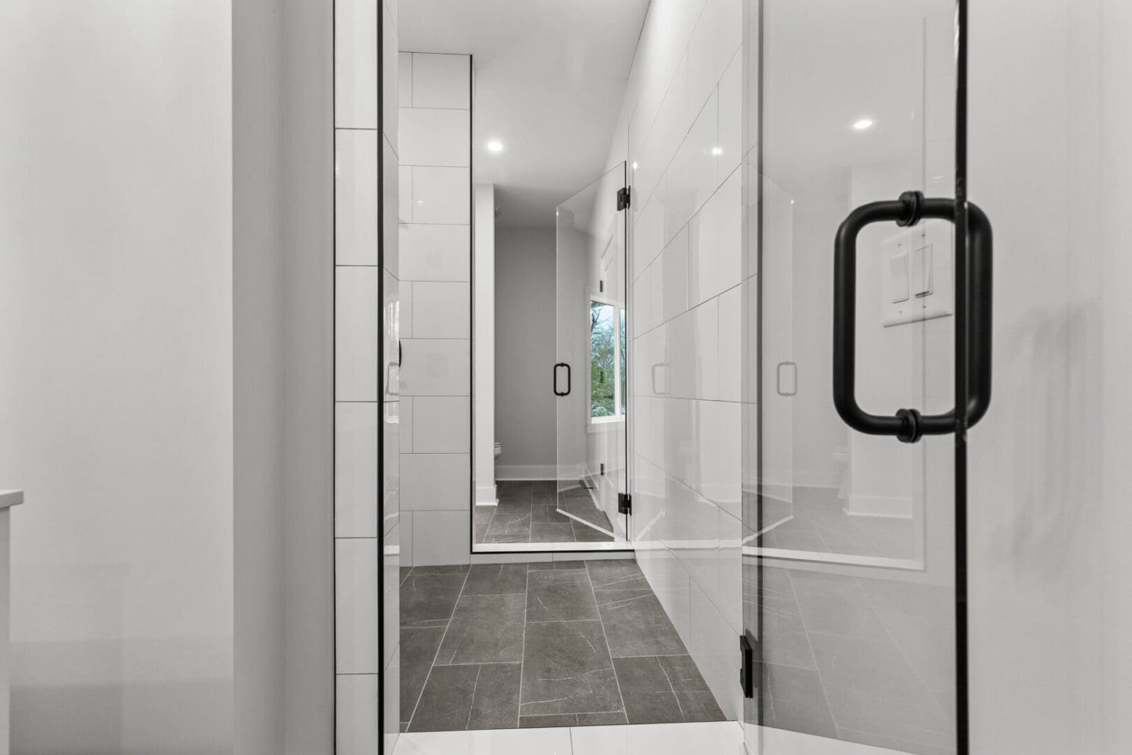 A modern bathroom featuring a glass shower enclosure with black handles, white tiled walls, and dark tiled floor. A large mirror and window are visible in the background.