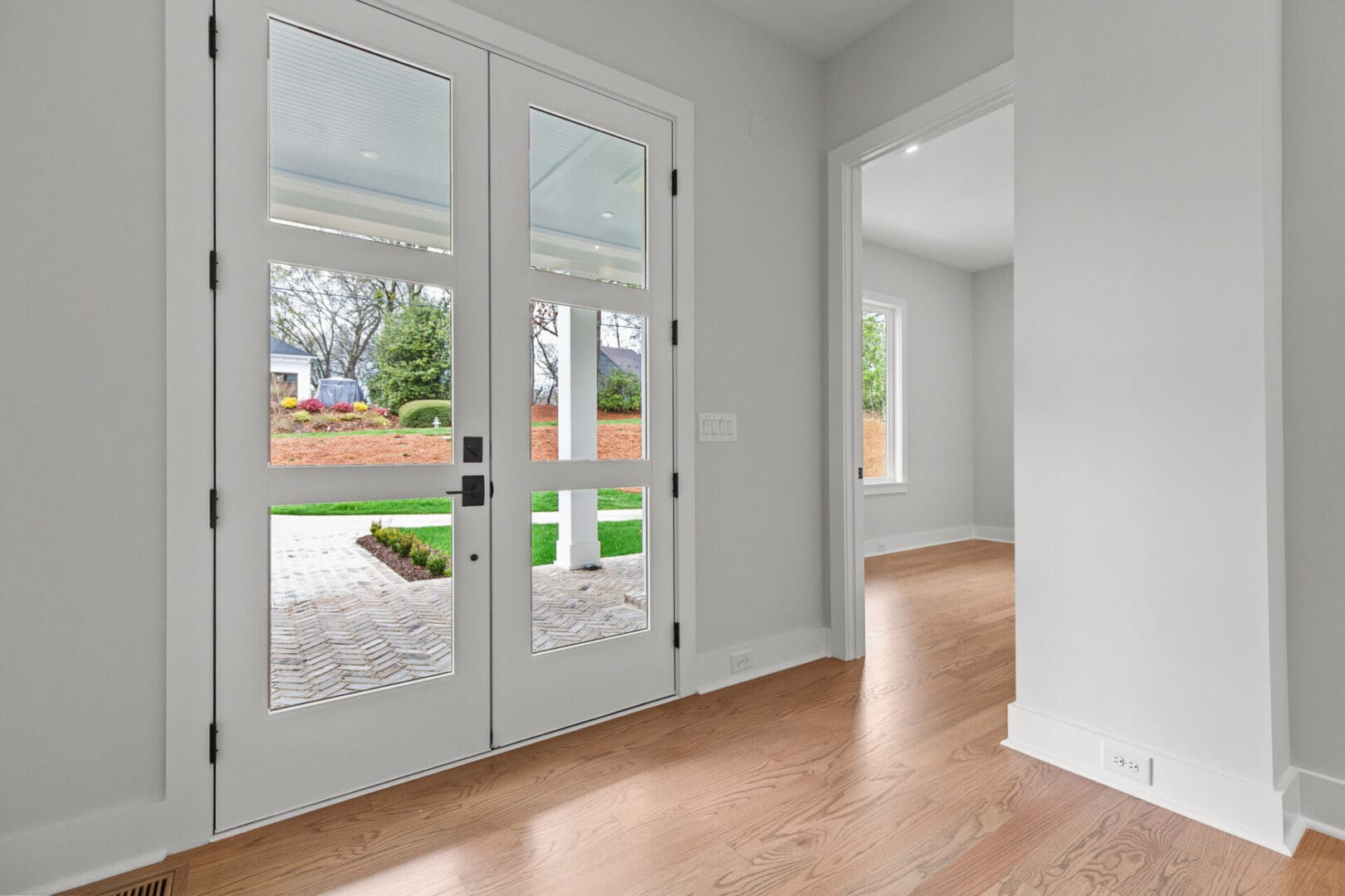 Interior view of a modern home featuring large glass double doors leading to a patio, with an adjacent room visible on the right. The flooring is light wood, and the walls are painted white.