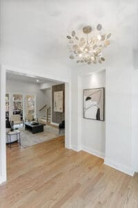 A modern interior with light wood floors, a white wall, abstract wall art, a decorative light fixture, and a view into a living room with black and white furnishings.