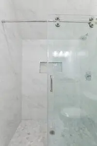 A modern shower with a sliding glass door, white marble tiles on the walls and floor, a small recessed shelf, and a chrome showerhead and fixtures.