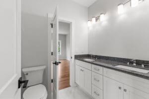 Modern bathroom with double sink vanity, black countertop, white cabinets, wall-mounted lights, toilet, and open door leading to adjacent room with wooden flooring.