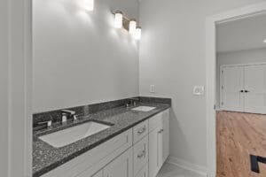 A modern bathroom features a double-sink vanity with dark granite countertops, white cabinets, and overhead lighting. The adjacent room has hardwood floors and white double doors.