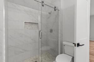 A modern bathroom featuring a glass-enclosed shower with marble tile and a built-in shelf, adjacent to a white toilet with a black handle on the flush lever.