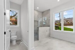A modern bathroom with a glass shower, tiled floors and walls, a toilet next to a window, and two large windows offering an outside view.