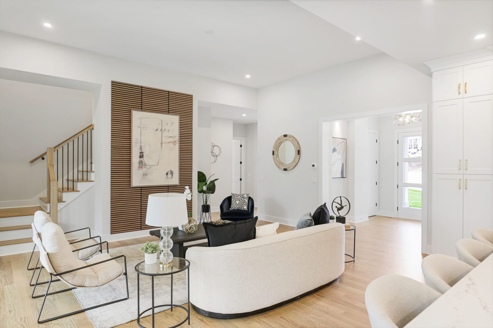 Modern living room with light wood flooring, white walls, and minimalist furniture, including a curved sofa, two armchairs, and a black accent chair. A staircase is visible to the left.