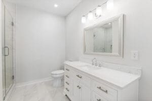 A well-lit bathroom with a white vanity, a large mirror, three light fixtures above, a toilet, and a glass-enclosed shower on the left. The decor is modern and minimalist.