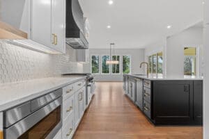 Modern kitchen with white cabinets, stainless steel appliances, black island, and hexagonal tile backsplash. Large windows provide ample natural light, and the hardwood floor accentuates the contemporary design.