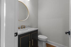 A modern bathroom with a round mirror, a marble countertop with dual faucets, and a black vanity cabinet. A toilet is positioned next to the cabinet against a light gray wall.