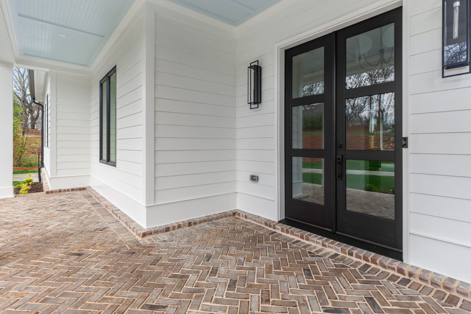 Front entrance of a building with large glass double doors, white siding, and a herringbone-patterned brick floor. The area is covered and features a minimalist, modern design.