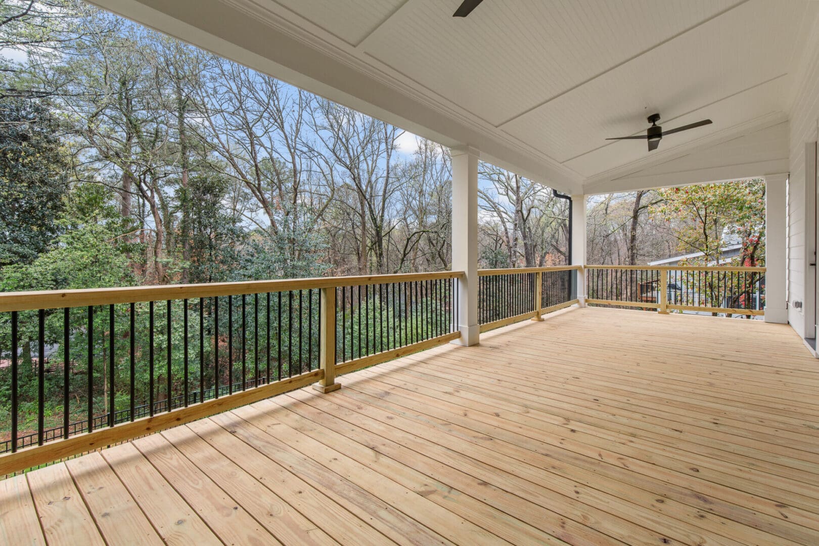 A spacious wooden deck with black railings overlooks a forested area. The deck has a ceiling fan and a white paneled roof.