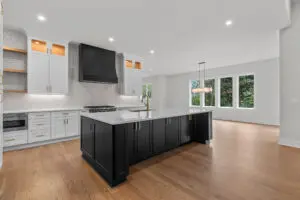 A spacious, modern kitchen with a large island featuring black cabinetry, white countertops, a black range hood, a built-in oven, and a contemporary light fixture.