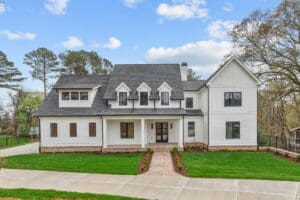 A two-story modern farmhouse with white siding, dark roof, and four dormer windows. It features a landscaped front yard with a brick path leading to a covered porch and front entrance.