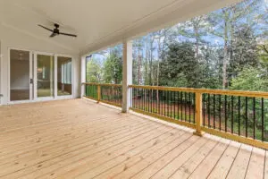 A spacious, covered wooden deck with a black ceiling fan, bordered by a railing, overlooks a lush, tree-filled backyard. Sliding glass doors lead into the house.