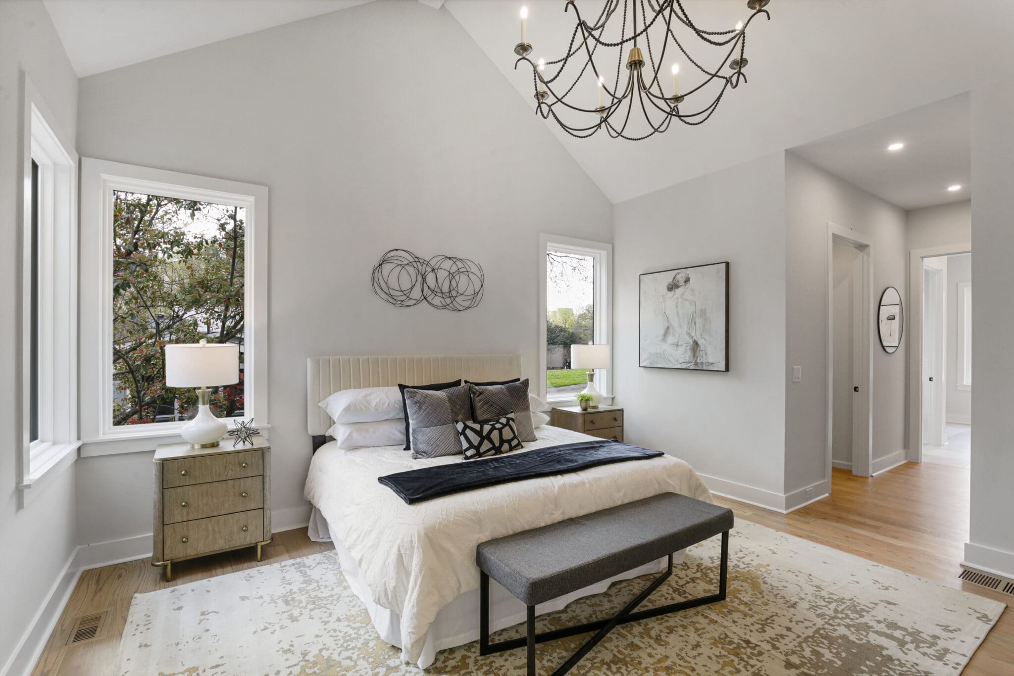 A modern bedroom with a large bed, gray bench, and two nightstands with lamps. The walls are light gray with a decorative wire art piece and a painting. There's a chandelier and natural light from windows.