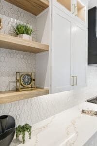 A modern kitchen features white hexagonal tile backsplash, a white countertop with marble veining, and wooden shelves holding a clock and plants. White cabinets and a darker cooking area are also visible.