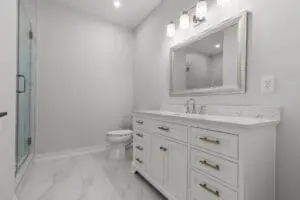 A modern bathroom featuring a white vanity with double sinks, a large framed mirror, a toilet, and a glass-enclosed shower with light-colored tiles.