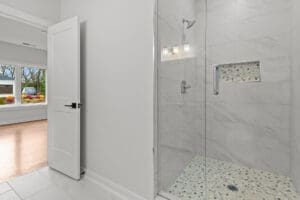 A modern bathroom featuring a glass-enclosed walk-in shower with a tiled wall and a small niche, located beside an open white door and an adjacent room with large windows and wooden floors.