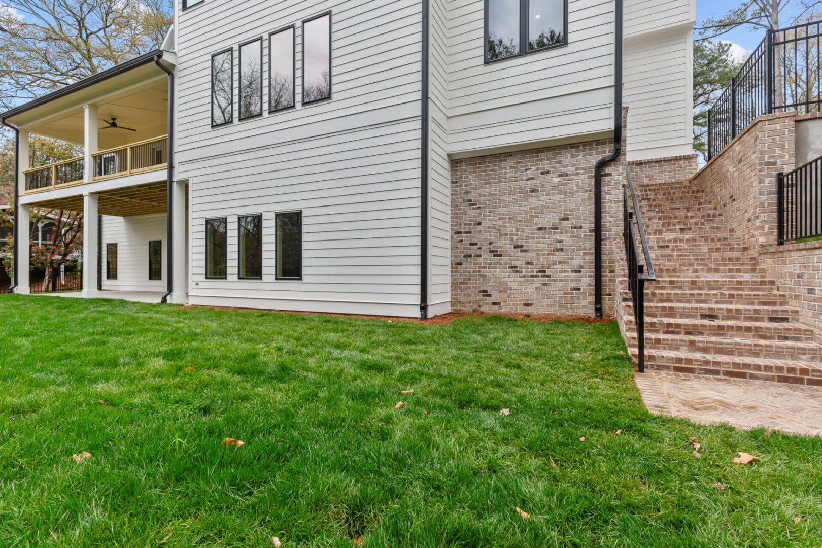 A modern white house with three large windows and a brick staircase on the right side, leading to an elevated deck. The house has a spacious grassy backyard.