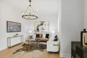 A modern living room with a brown sofa, white armchairs, a cowhide rug, and abstract wall art. A black chandelier hangs from the ceiling, and a console table with decor is against one wall.