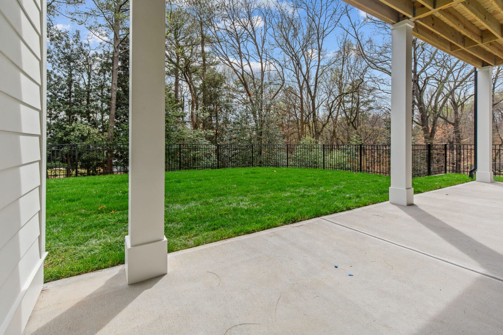A covered concrete patio opens to a fenced green lawn, bordered by trees and shrubs under a clear sky.