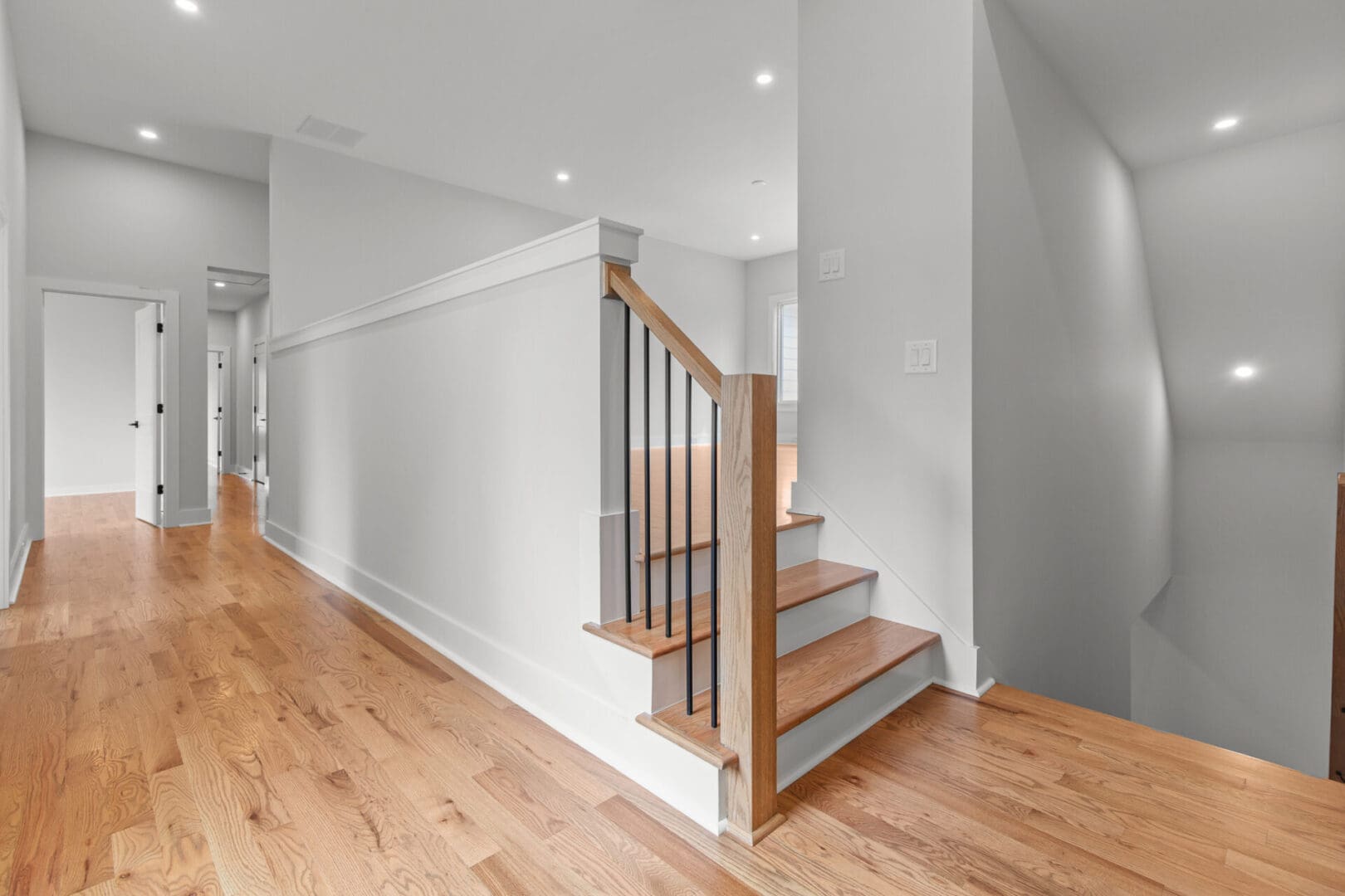 A brightly lit hallway with wooden floors, white walls, and a staircase with a wooden banister and black balusters. There are doors along the hallway and a light fixture on the ceiling.