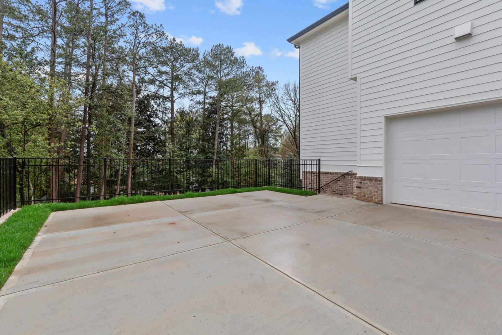 A concrete driveway leads to a white garage attached to a house, with a metal fence and trees in the background under a blue sky.