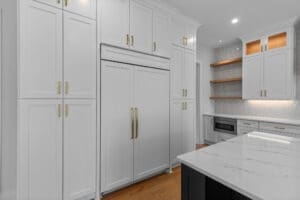 Modern kitchen with white cabinets, an integrated refrigerator, and gold handles. There are floating wooden shelves, a built-in microwave, and a marble countertop island.