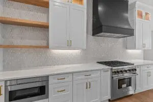 A modern kitchen features white cabinetry with gold handles, hexagon-tiled backsplash, open wooden shelves, stainless steel appliances, and a black hood over a gas stove.