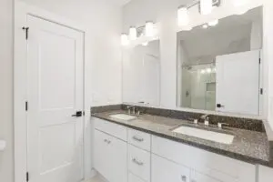 A modern bathroom with white cabinets, a granite countertop, dual sinks, a large mirror, and bright lighting. The room also features a door and a glass-enclosed shower.