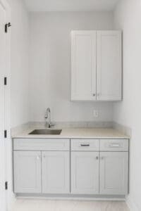 A small, modern kitchen alcove with gray cabinets, a countertop, a stainless steel sink, and a wall-mounted cabinet above. The walls are white and minimalistic.