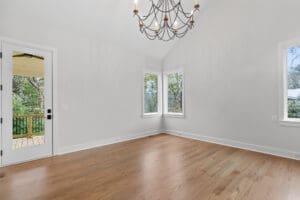 An empty room with wooden floors, white walls, a chandelier, three windows with outdoor views, and a glass door leading to a wooden deck.